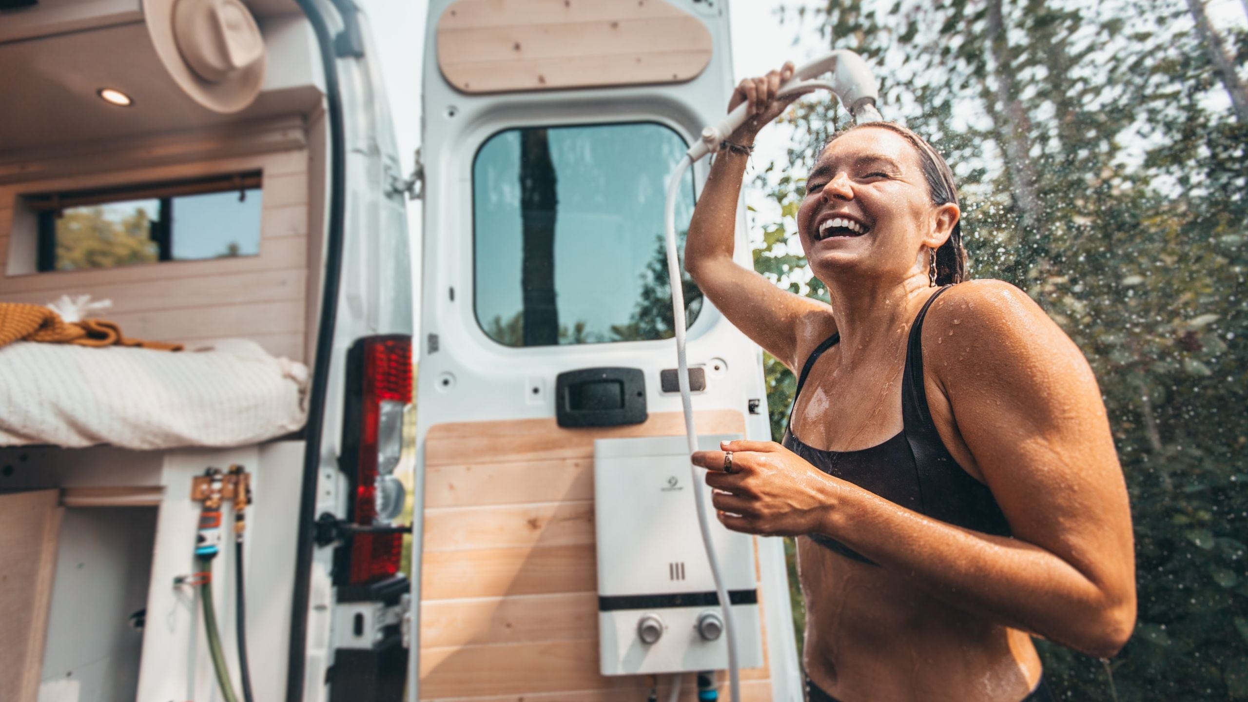Smiling woman taking an outdoor shower next to a camper van in a forest setting.