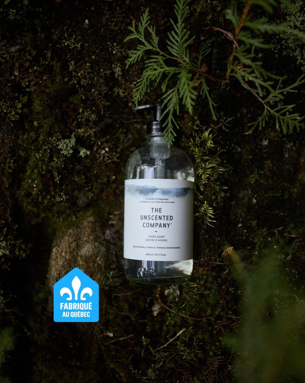 Bottle of The Unscented Company hand soap placed upright against a mossy rock wall, with forest greenery. A “Fabriqué au Québec” badge appears in the lower left.