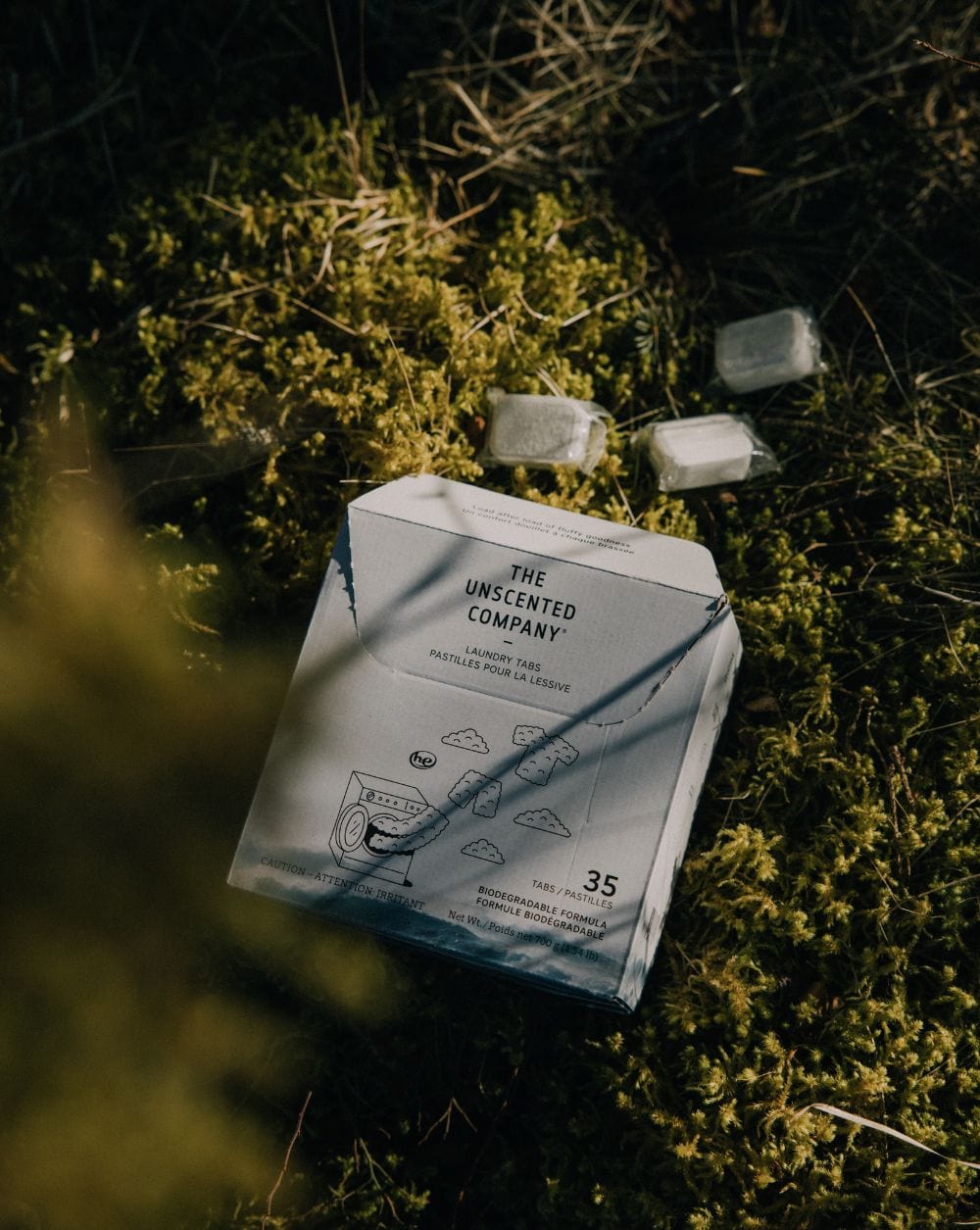 Box of The Unscented Company laundry tabs placed on mossy ground, with four white laundry tablets scattered nearby. The box shows “35 tabs” and “biodegradable formula.”