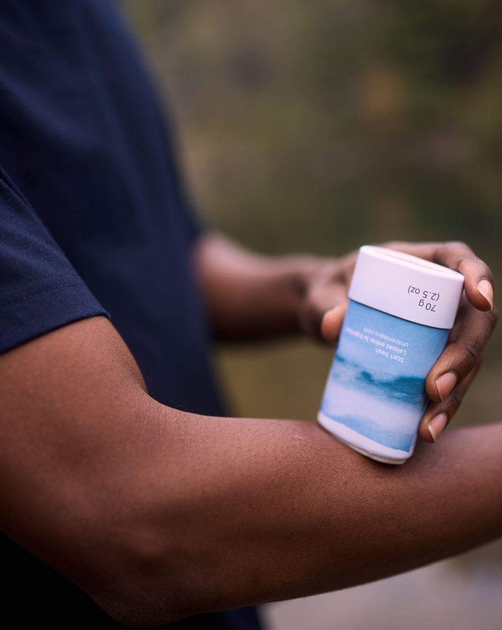 A person in a dark t-shirt holds a deodorant stick and applies it to their forearm. The deodorant has a blue and white label.
