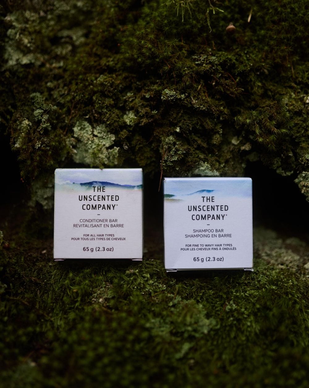 Two boxes of The Unscented Company bars, one conditioner and one shampoo, placed on green moss outdoors.