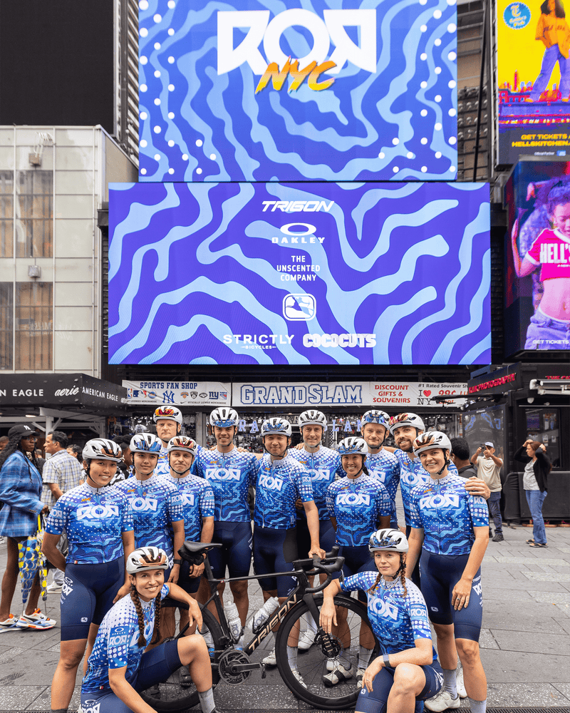 Cycling team posing in Times Square beneath a large digital billboard featuring sponsors including The Unscented Company, highlighting community partnerships and urban cycling culture.