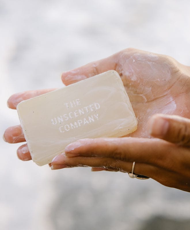 Close-up of hands holding a bar of soap with "The Unscented Company" logo.