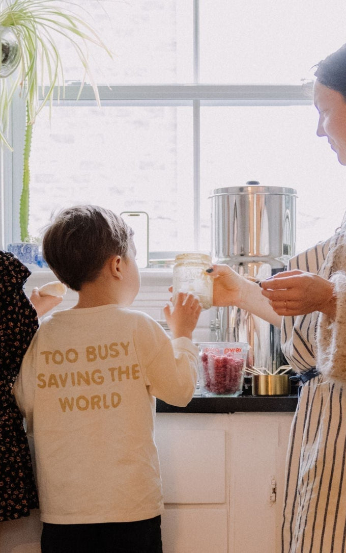 A woman and three young children preparing food together in a bright kitchen. One child wears a shirt with the words 'Too busy saving the world' while the woman holds a jar of ingredients near a sunny window.