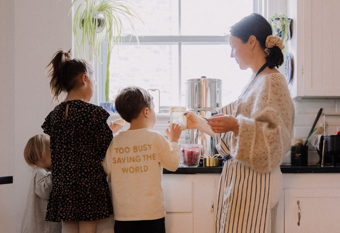 A woman and three young children preparing food together in a bright kitchen. One child wears a shirt with the words 'Too busy saving the world' while the woman holds a jar of ingredients near a sunny window.