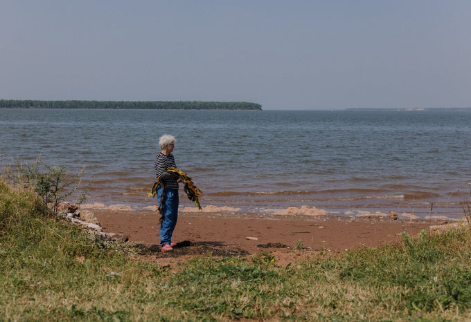 An elderly woman with white hair, wearing a striped shirt and blue pants, stands on a beach holding freshly harvested seaweed, with the ocean and distant forested shoreline in the background.
