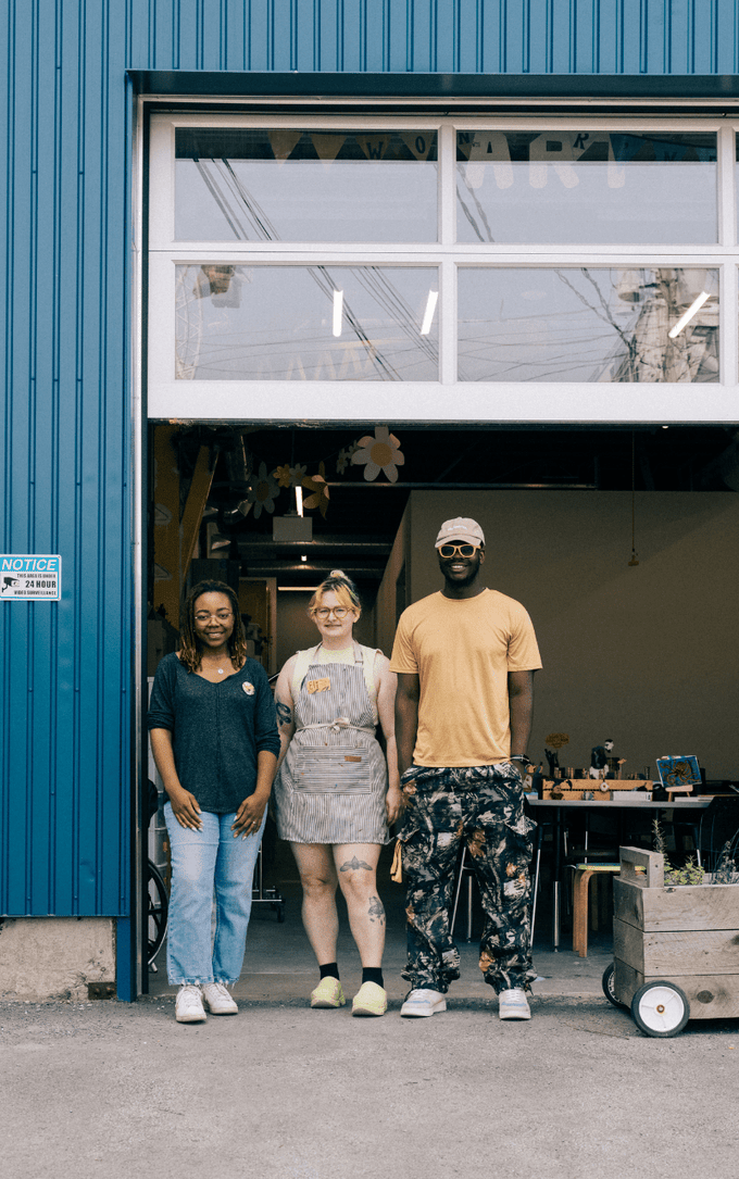 Three people standing in front of an open garage-style workshop door of a blue industrial building. A bicycle with a green crate is parked on the left, while a wooden planter box sits on the right. Inside, worktables and shelves with supplies are visible.