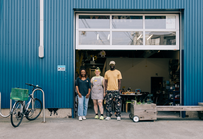 Three people standing in front of an open garage-style workshop door of a blue industrial building. A bicycle with a green crate is parked on the left, while a wooden planter box sits on the right. Inside, worktables and shelves with supplies are visible.