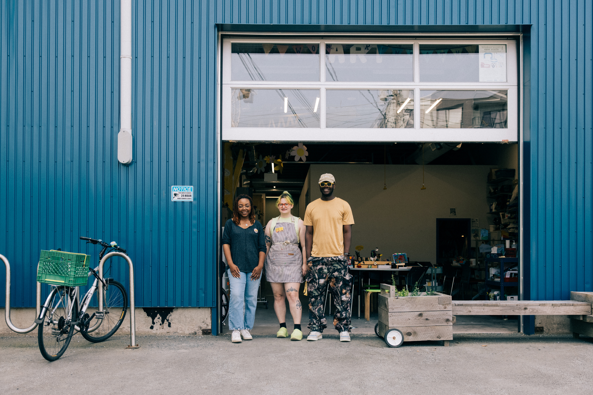 Three people standing in front of an open garage-style workshop door of a blue industrial building. A bicycle with a green crate is parked on the left, while a wooden planter box sits on the right. Inside, worktables and shelves with supplies are visible.