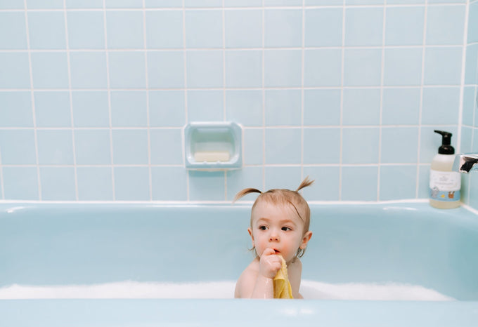 A toddler with small pigtails sitting in a blue bubble bath, holding a yellow washcloth up to their mouth, with a bottle of The Unscented Company Gentle Baby Wash & Shampoo sitting on the edge of the tub.