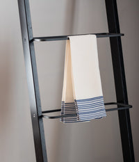 A cream-colored textured towel with navy blue stripes hanging neatly on a black metal ladder against a neutral wall.