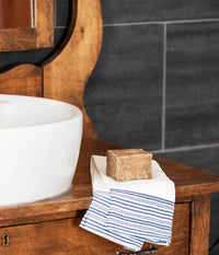 A rustic brown soap bar resting on a striped cream towel next to a white ceramic sink on a wooden vanity, with dark tile wall in the background.
