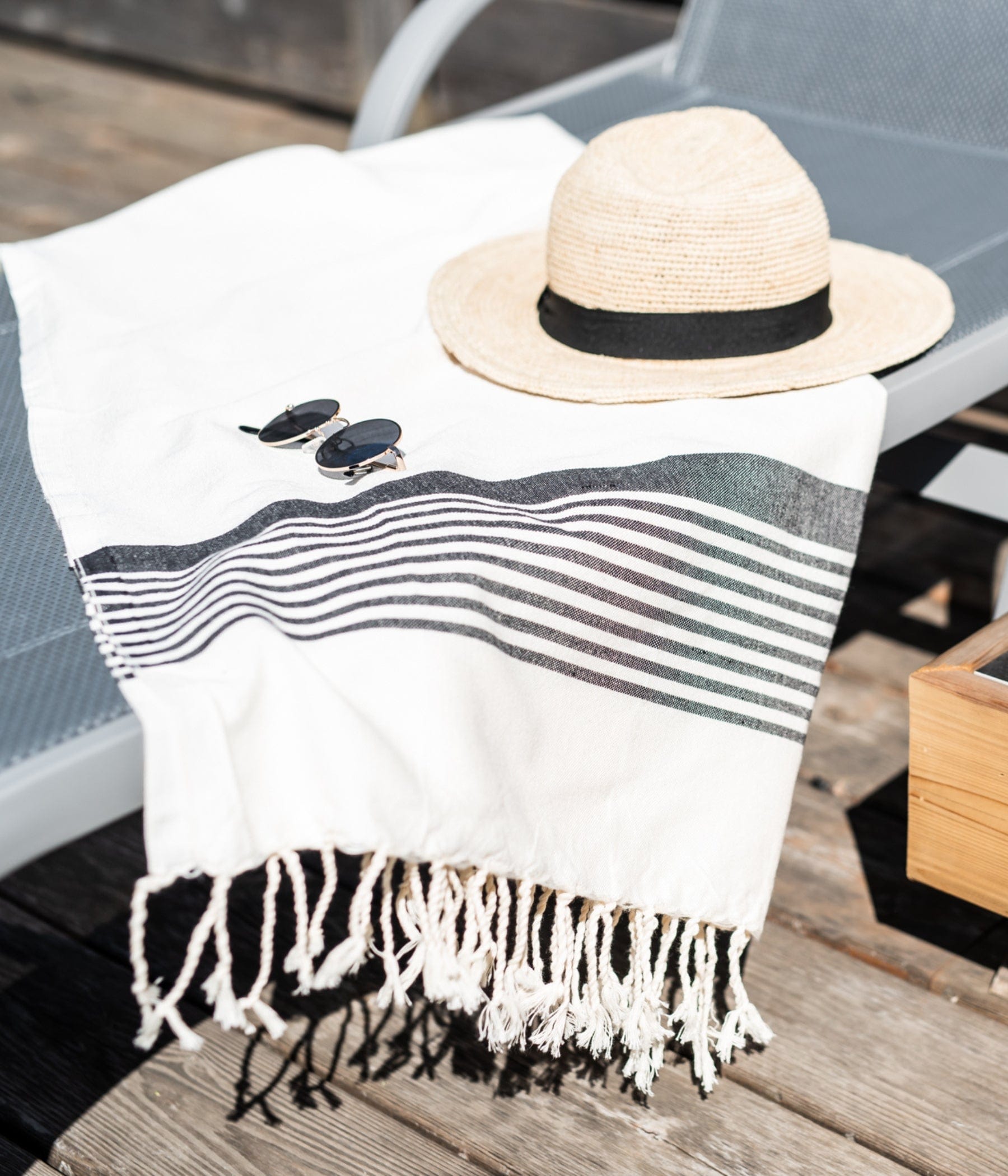 A straw hat and sunglasses resting on a striped fringed towel draped over a lounge chair on a wooden deck, suggesting a relaxed summer atmosphere.
