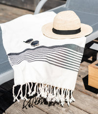 A straw hat and sunglasses resting on a striped fringed towel draped over a lounge chair on a wooden deck, suggesting a relaxed summer atmosphere.