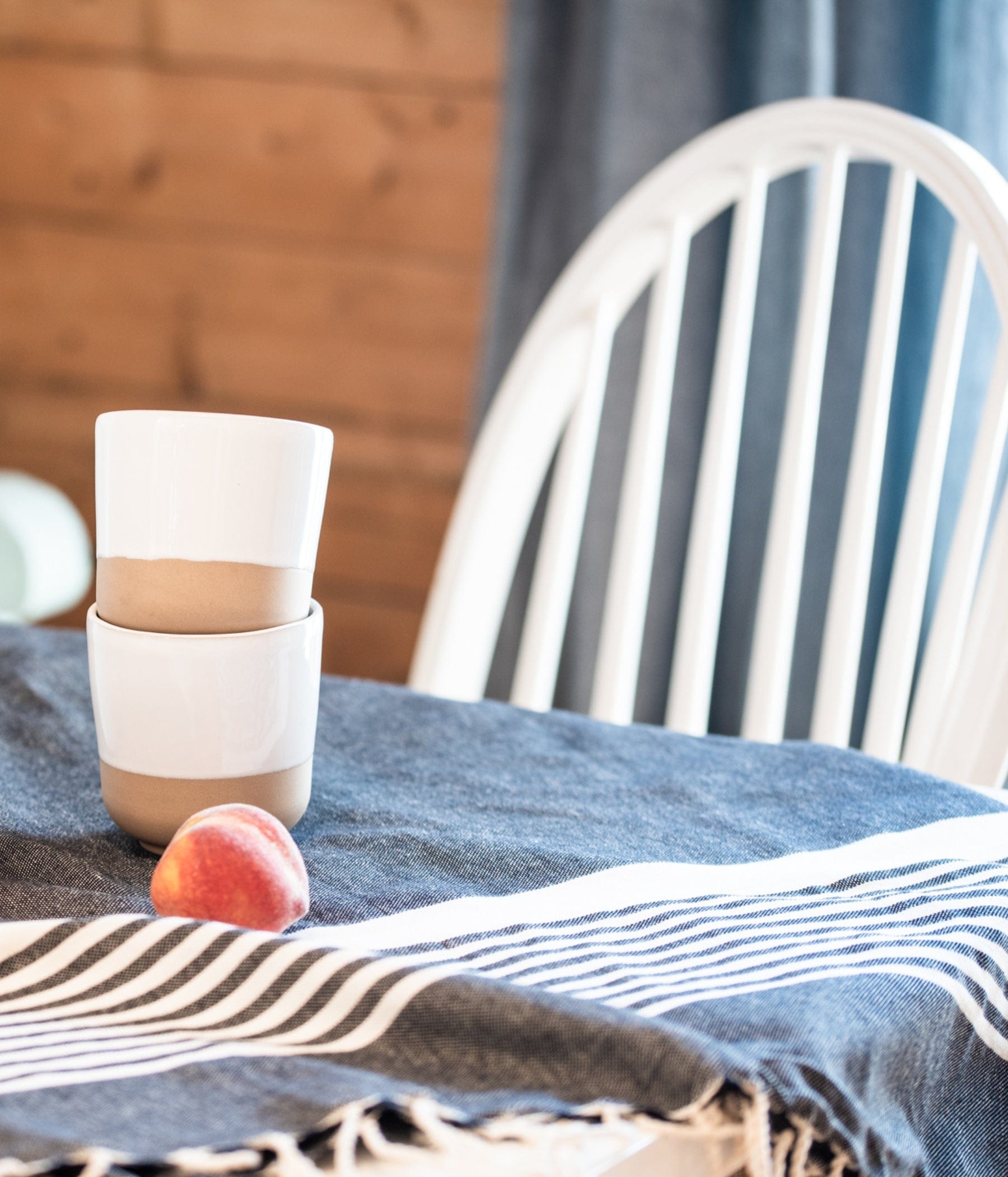Two stacked ceramic cups and a fresh peach placed on a striped cloth over a table, with a white wooden chair and cozy interior in the background.