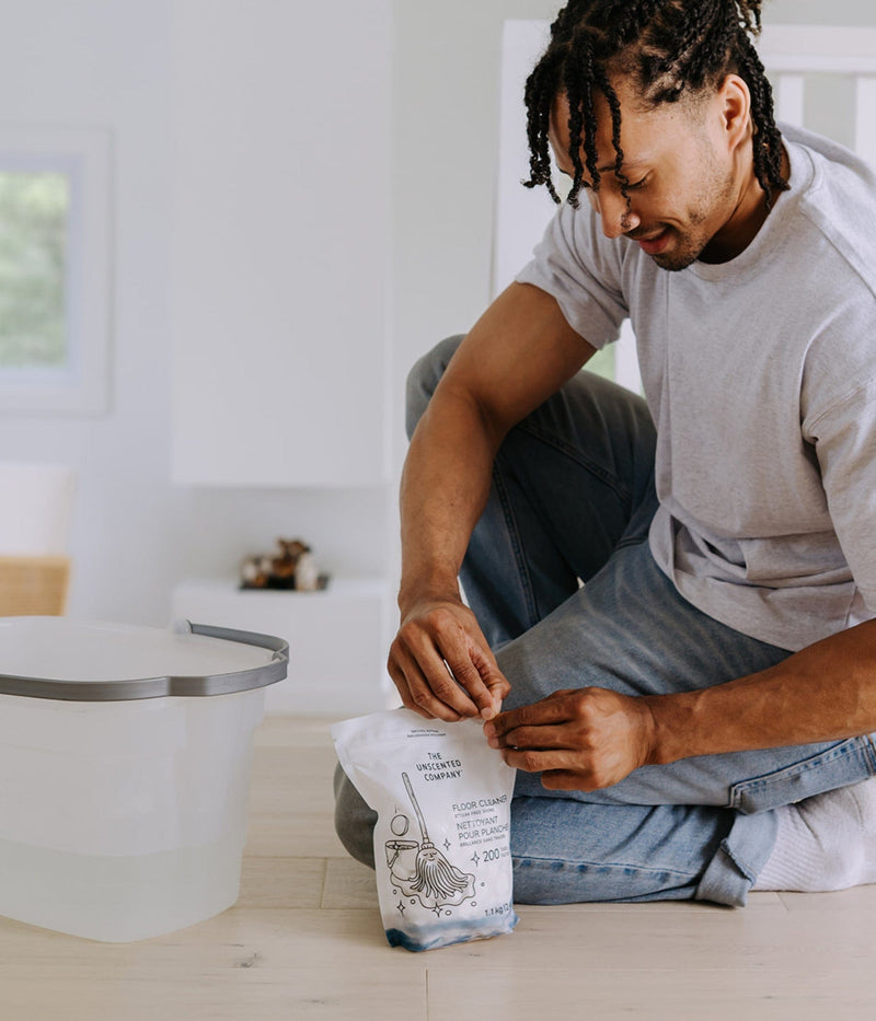 A man kneels on a light wood floor beside a bucket of water, opening a pouch of The Unscented Company Floor Cleaner tablets. He is wearing a grey t-shirt and jeans, preparing to clean.