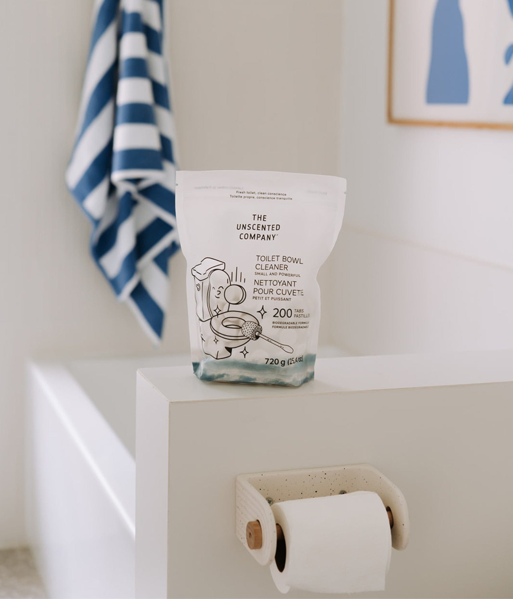 A pack of The Unscented Company Toilet Bowl Cleaner (200 tabs) placed on the edge of a white bathtub. In the background, a blue and white striped towel hangs beside a framed minimalist artwork.