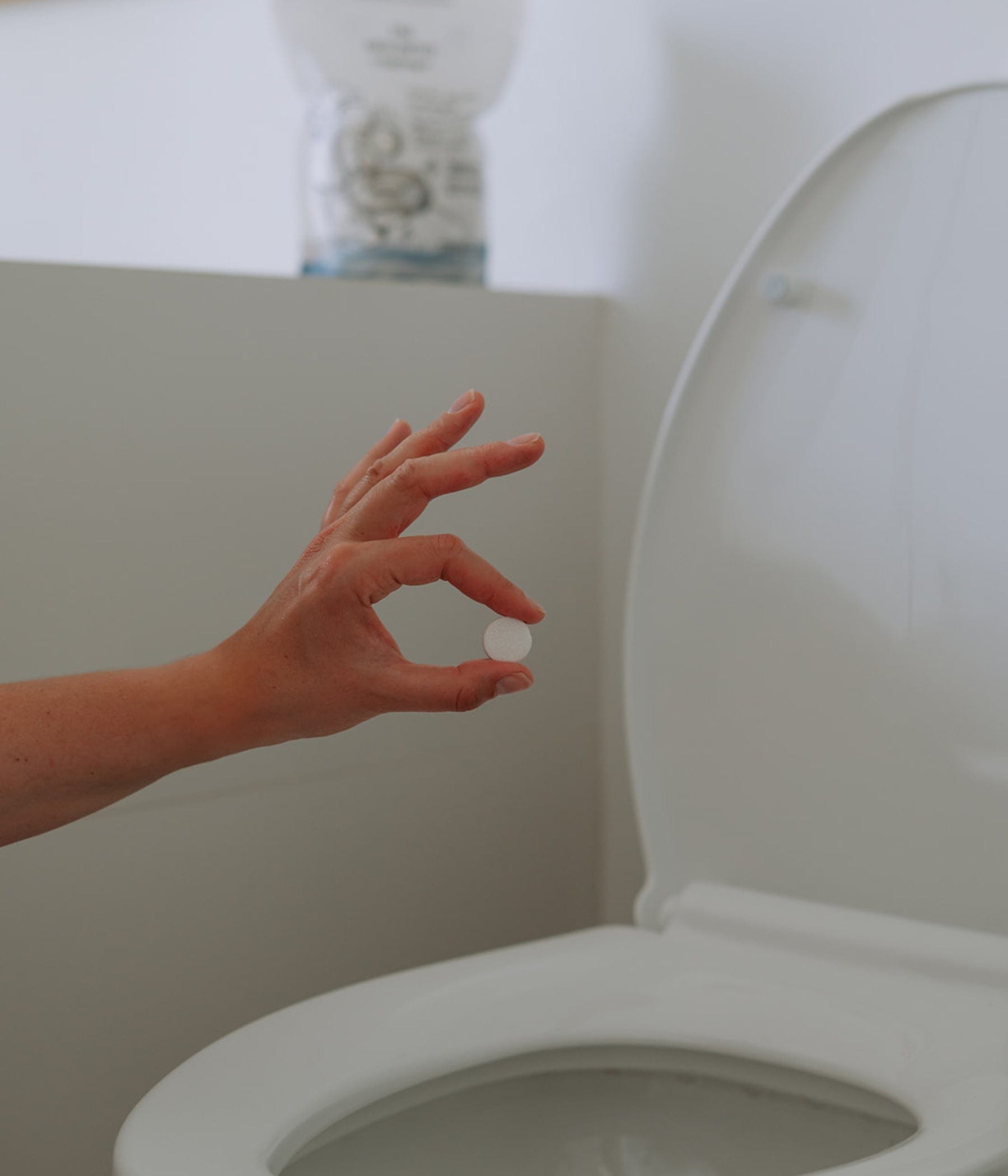 A close-up of a hand holding a single toilet bowl cleaning tablet above an open toilet bowl. The product pouch is blurred in the background on the bathtub edge.