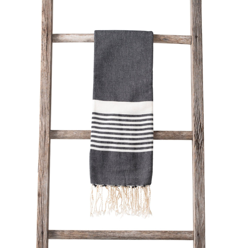 A dark grey towel with white and black stripes and fringe ends, neatly draped over a rustic wooden ladder.