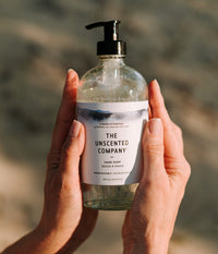 A bottle of The Unscented Company hand soap held with both hands in soft light.