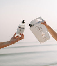 Two hands holding a hand soap bottle and refill pouch from The Unscented Company in front of the sea.