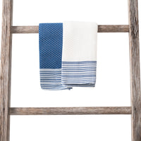 Two folded towels hanging side by side on a wooden ladder, one blue with a textured pattern and stripes, the other cream with matching blue-striped ends.