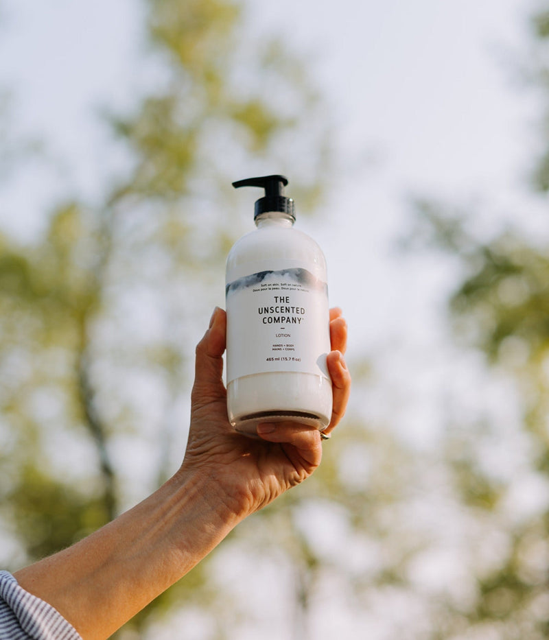 A hand lifts a bottle of The Unscented Company lotion against a blurry tree background.