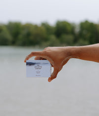 A sandy hand presents a bar of The Unscented Company soap in front of a body of water.