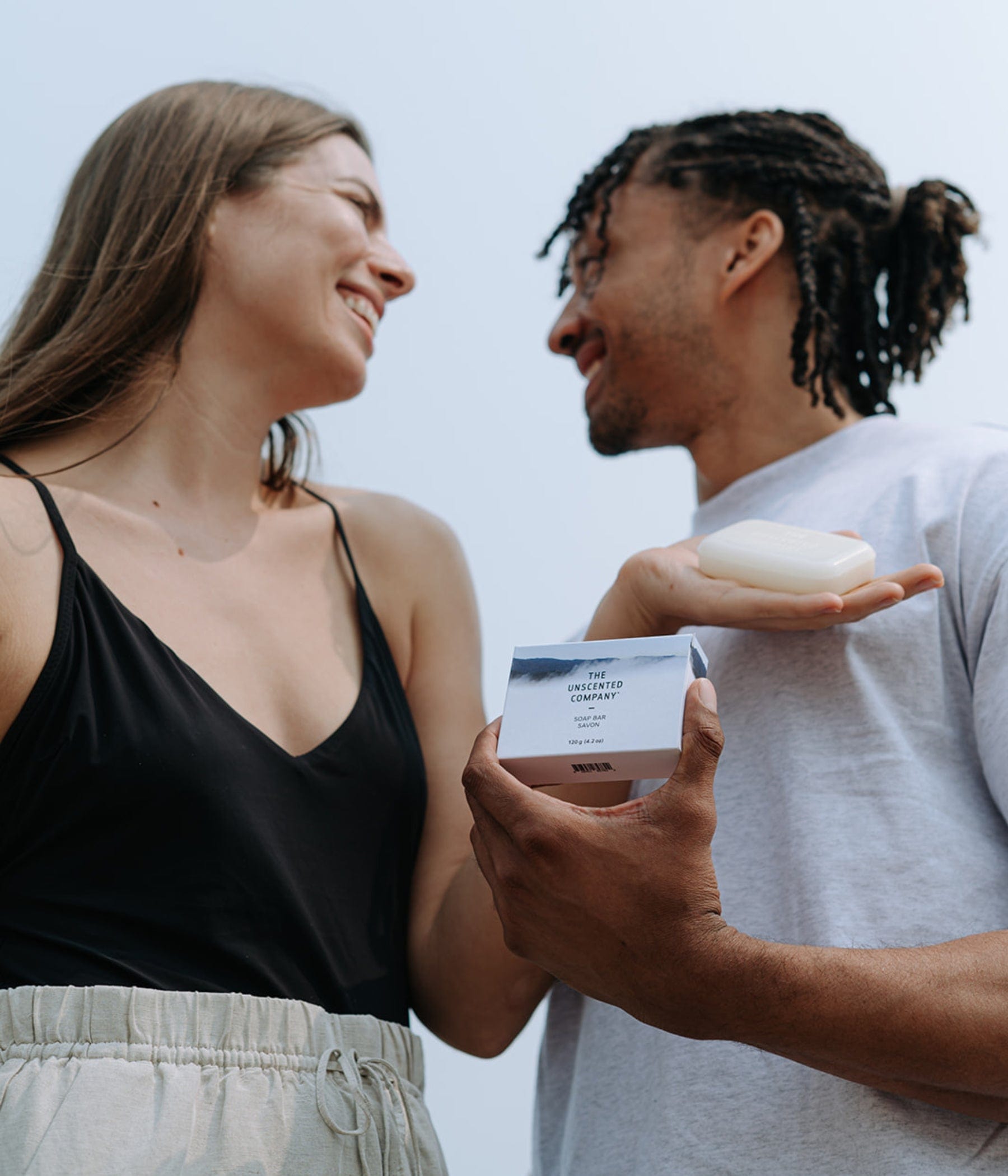 A smiling couple looks at each other while holding a bar of The Unscented Company soap.