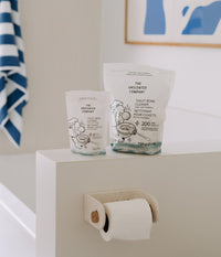 Two pouches of The Unscented Company toilet bowl cleaner, one small (22 tabs) and one large (200 tabs), placed on the edge of a bathtub near a toilet paper roll.