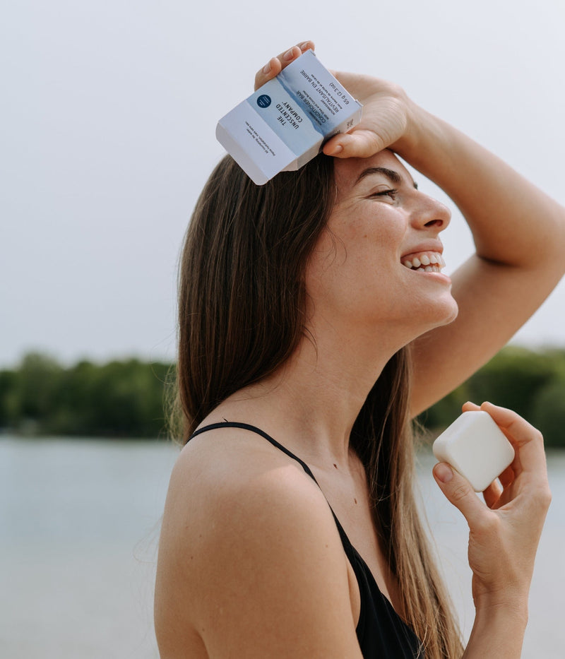 Smiling woman holding The Unscented Company shampoo bar and box near her head.