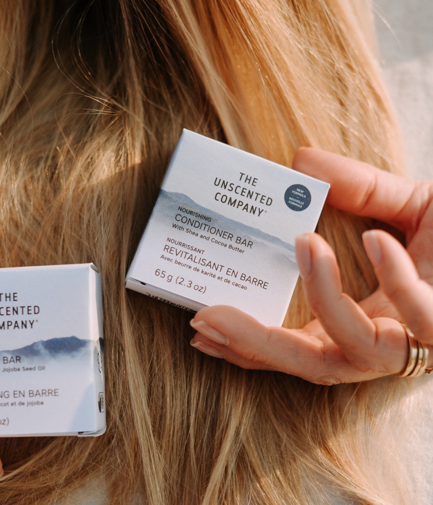 The Unscented Company conditioner bar resting in a woman’s hand on her long blonde hair.