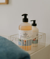 The Unscented Company Shiny Shampoo + Wash for kids in a reusable pump bottle, displayed in a white wire basket in a bright bathroom.
