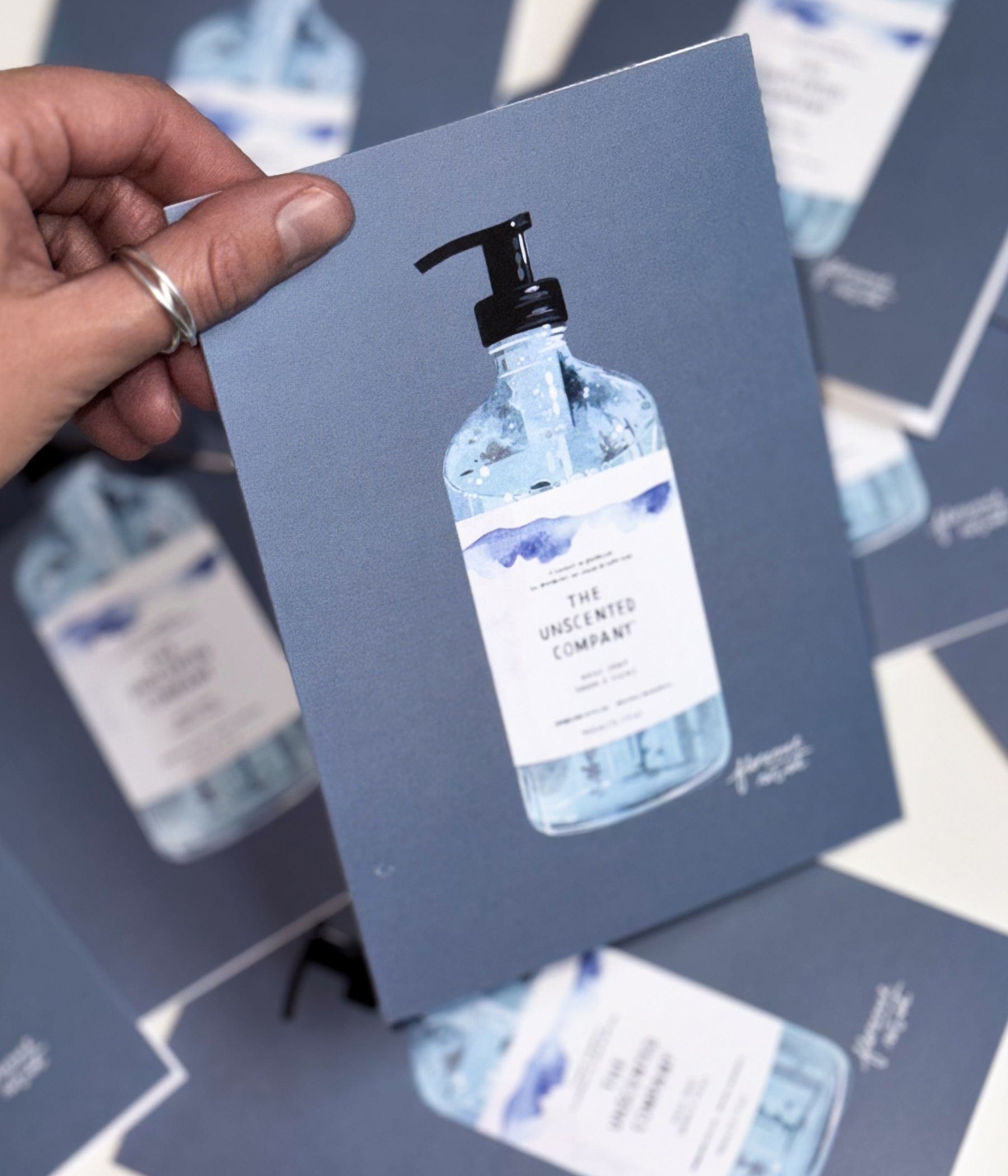 Vue rapprochée d’une carte de souhaits illustrée représentant une bouteille de savon pour les mains de The Unscented Company, tenue par une personne, avec plusieurs cartes similaires étalées en arrière-plan.