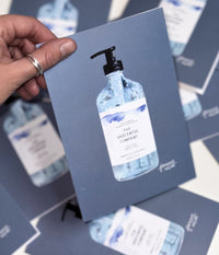 Vue rapprochée d’une carte de souhaits illustrée représentant une bouteille de savon pour les mains de The Unscented Company, tenue par une personne, avec plusieurs cartes similaires étalées en arrière-plan.
