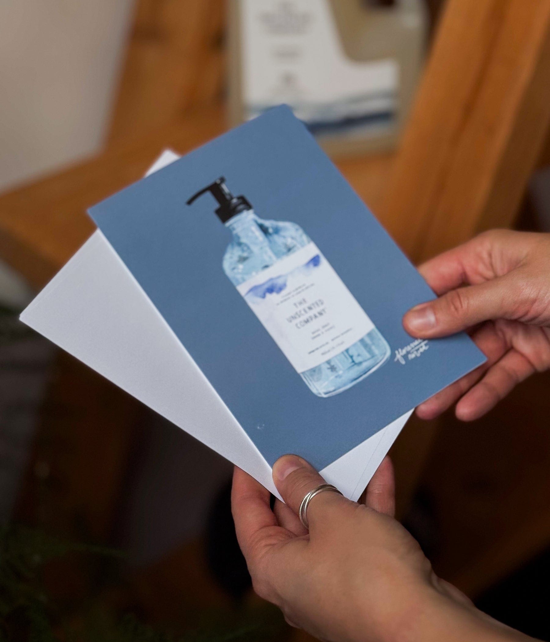 Carte de souhaits illustrée montrant une bouteille de savon pour les mains de The Unscented Company, tenue entre les mains d’une personne, avec une enveloppe blanche en arrière-plan.