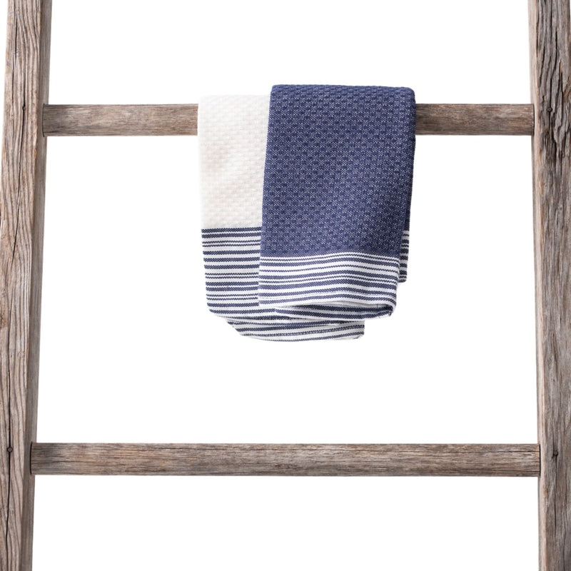 A pair of cotton towels in cream and navy tones with woven textures and striped edges, neatly displayed on a wooden ladder.