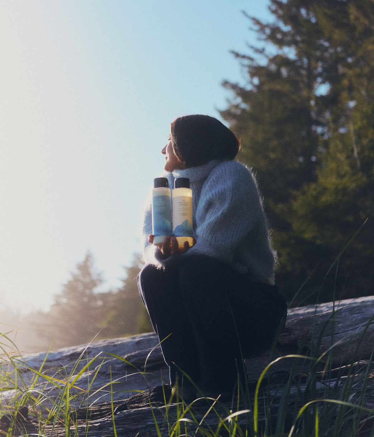 A woman wearing a soft light-blue turtleneck sweater and dark pants sits outdoors on a large fallen tree near a coastal forest, holding two 473 mL bottles of The Unscented Company volumizing shampoo and conditioner. The white hair care bottles feature black caps and blue botanical graphics, illuminated by warm natural sunlight with tall evergreen trees, beach grass, and soft coastal mist in the background.