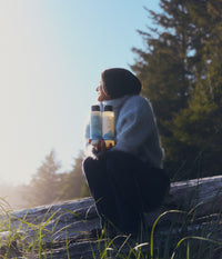 A woman wearing a soft light-blue turtleneck sweater and dark pants sits outdoors on a large fallen tree near a coastal forest, holding two 473 mL bottles of The Unscented Company volumizing shampoo and conditioner. The white hair care bottles feature black caps and blue botanical graphics, illuminated by warm natural sunlight with tall evergreen trees, beach grass, and soft coastal mist in the background.