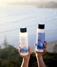 Two hands hold 473 mL bottles of The Unscented Company Volumizing Shampoo and Nourishing Conditioner against a sunlit coastal landscape. The unscented hair care bottles feature black caps and minimalist white-and-blue packaging, with ocean waves, sandy beach, and coastal greenery softly blurred in the background.