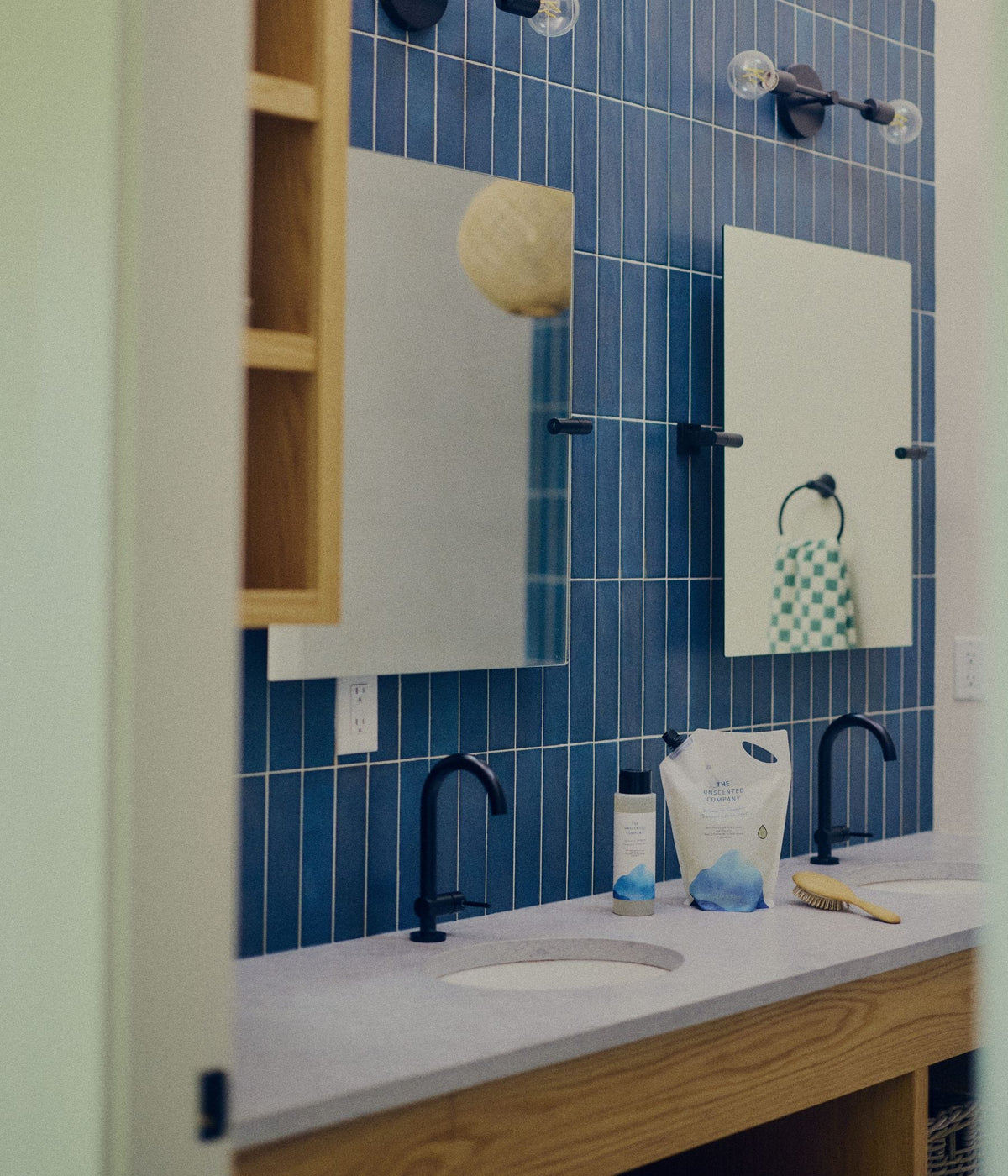 A modern bathroom vanity with a white countertop, two sinks, matte black faucets, rectangular mirrors, and a deep blue tiled wall. A 473 mL bottle and a large refill pouch of The Unscented Company volumizing hair care products sit beside a wooden hairbrush on the counter, highlighting the brand’s unscented shampoo and refillable hair care packaging in a clean bathroom setting.