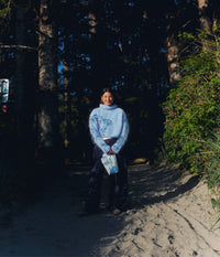 A woman wearing a soft light-blue turtleneck sweater and dark pants stands on a sandy forest path, smiling toward the camera while holding a white and blue The Unscented Company hair care refill pouch. She is surrounded by tall trees and dense green vegetation, with bright natural sunlight creating strong shadows across the coastal forest setting.
