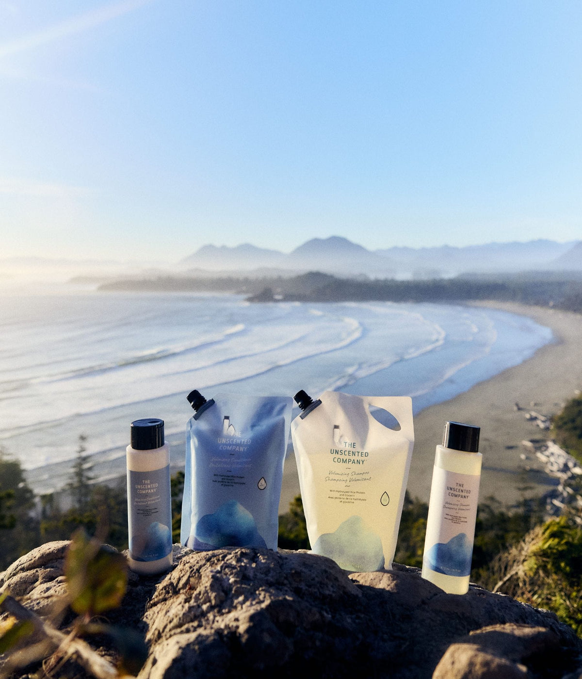 Four The Unscented Company hair care products—including two 473 mL bottles and two refill pouches for volumizing shampoo and conditioner—are arranged on a rocky overlook. The unscented hair care products feature white and blue packaging and are photographed in natural sunlight above a sweeping coastal beach, ocean waves, forest, and distant mountains.