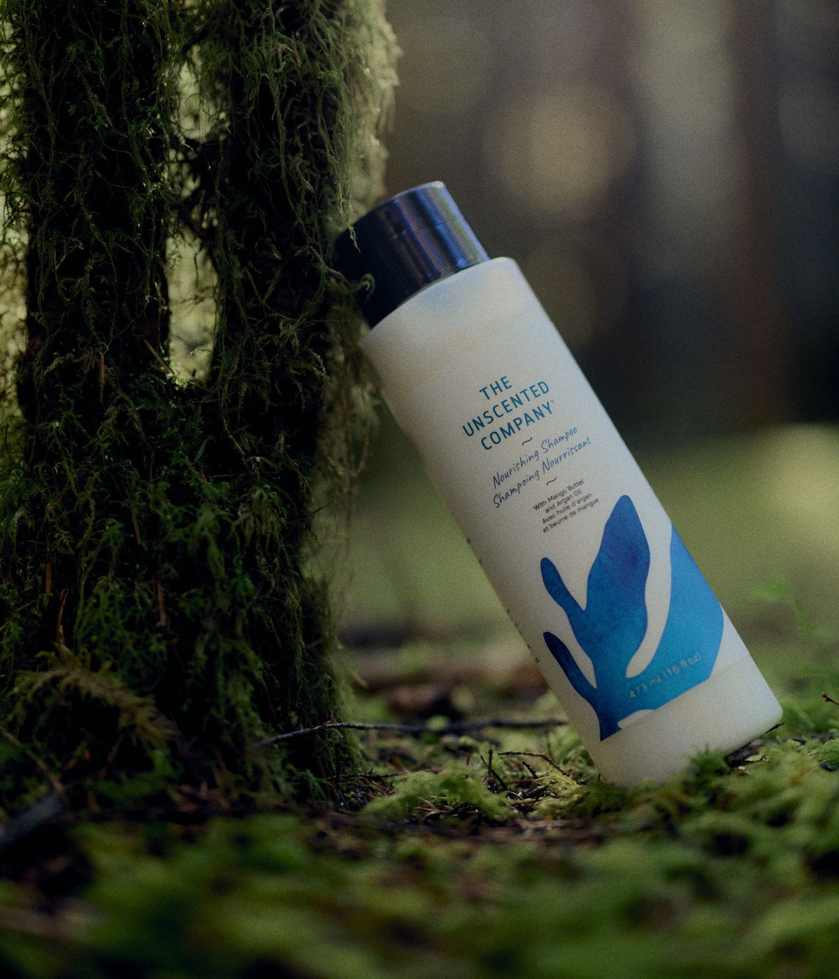 A 473 mL bottle of The Unscented Company nourishing shampoo with blue botanical graphics leans against a moss-covered tree on a green forest floor. The hair care product is formulated with mango butter and argan oil and is photographed among moss, branches, and soft natural woodland light.