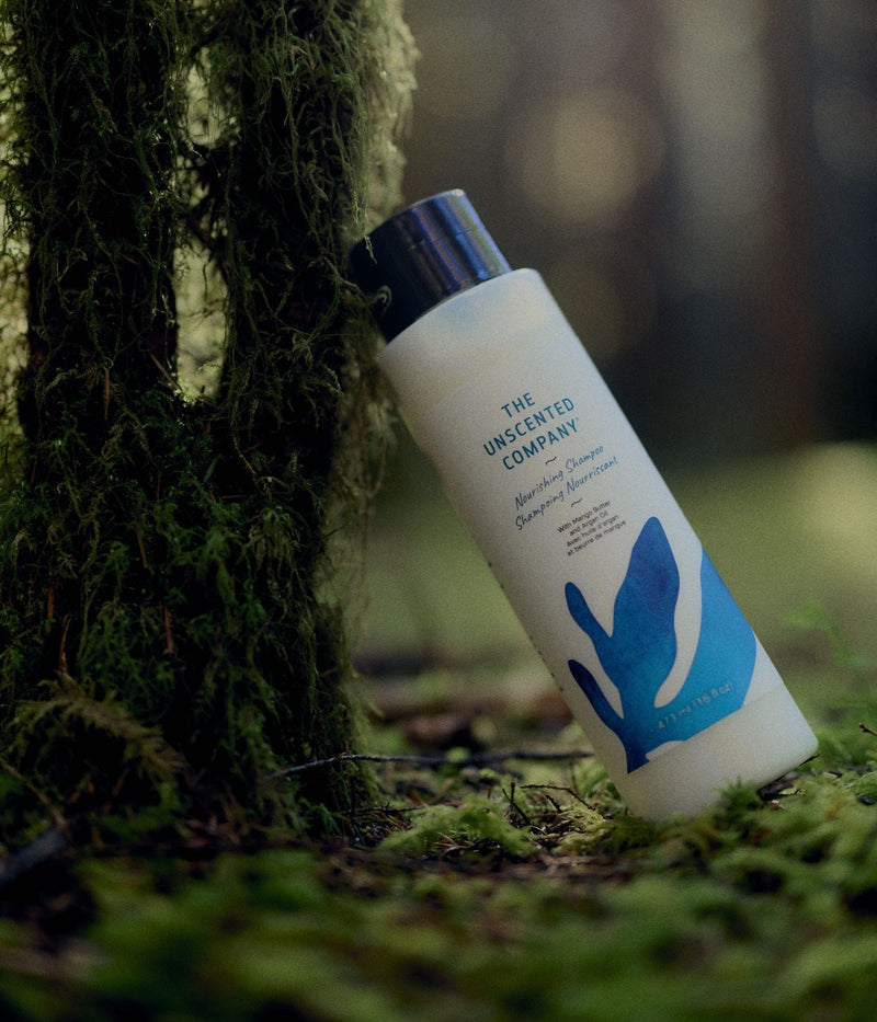 A 473 mL bottle of The Unscented Company nourishing shampoo with blue botanical graphics leans against a moss-covered tree on a green forest floor. The hair care product is formulated with mango butter and argan oil and is photographed among moss, branches, and soft natural woodland light.