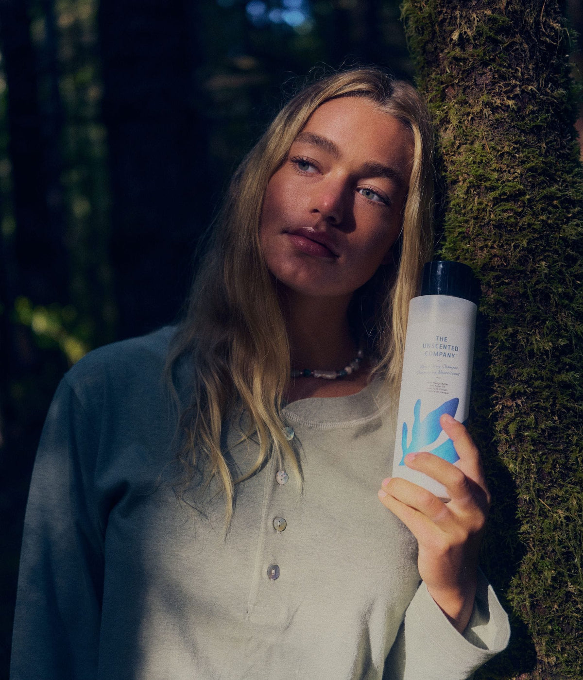 A blonde woman wearing a light sage-green long-sleeve top stands beside a moss-covered tree in a shaded forest, holding a bottle of The Unscented Company nourishing shampoo with blue botanical graphics, formulated with mango butter and argan oil, as sunlight falls across her face and the product.
