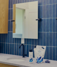 A modern bathroom countertop with a white sink, matte black faucet, blue tiled wall, and rectangular mirror. Beside the sink are The Unscented Company nourishing shampoo bottle and 2-litre refill pouch with blue botanical graphics, made with mango butter and argan oil, alongside a small blue-and-white patterned hair clip.