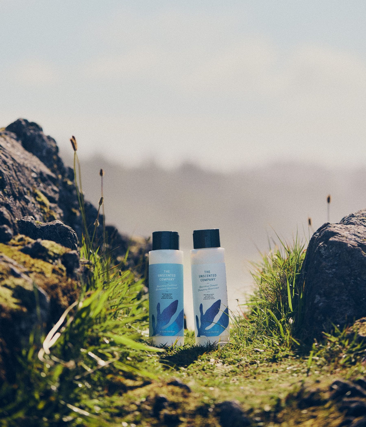 Bottles of The Unscented Company nourishing shampoo and nourishing conditioner with mango butter and argan oil stand together among mossy rocks and green grass in a sunlit mountain landscape.