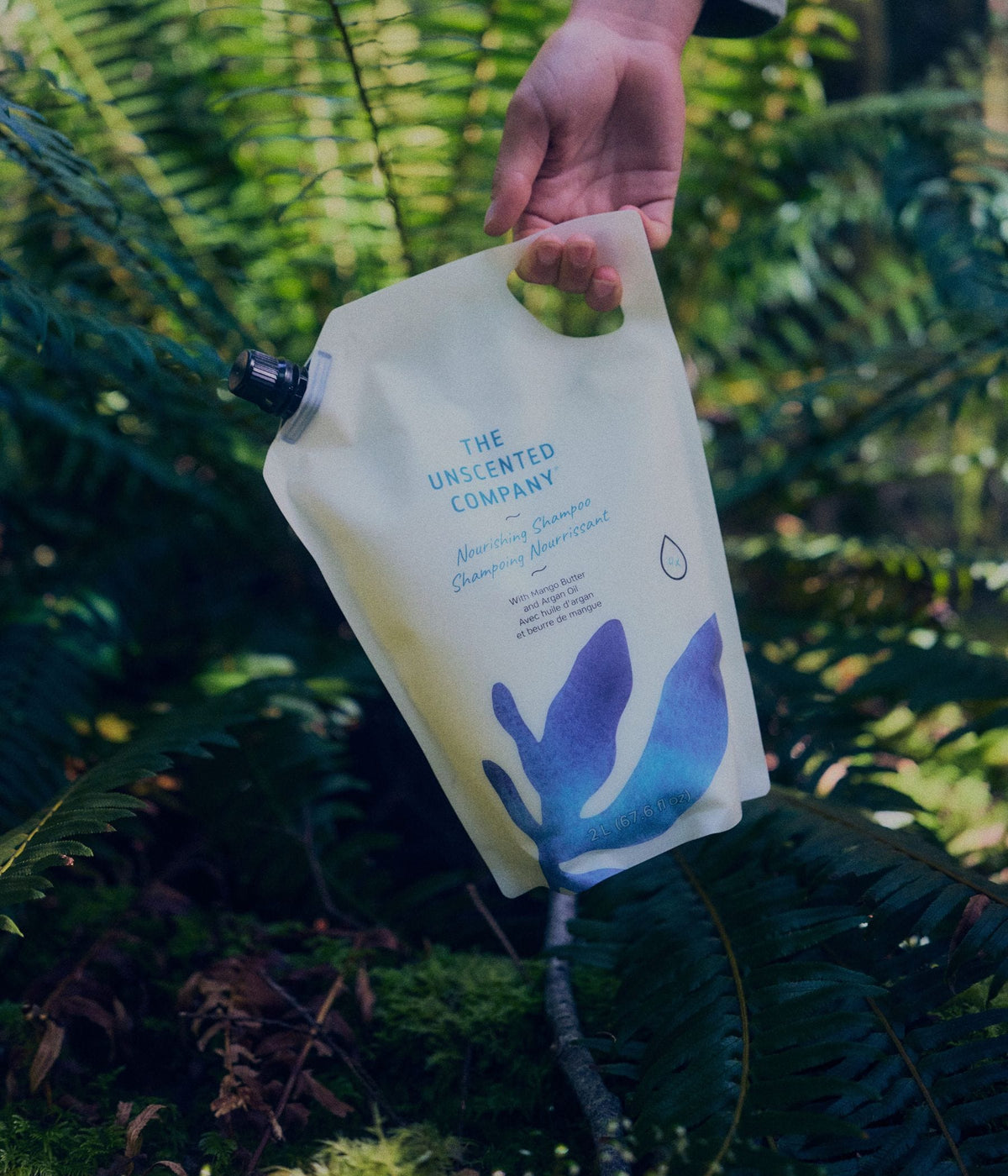 A hand holds a 2-litre refill pouch of The Unscented Company nourishing shampoo with mango butter and argan oil among green ferns and moss on a forest floor.