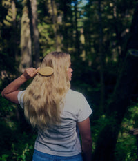 A blonde woman in a white T-shirt and blue jeans, seen from behind, brushes her long wavy hair with a wooden hairbrush while standing in a sunlit green forest.